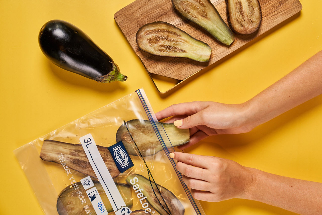 Eine Person legt gebratene Auberginenscheiben in einen SafeLoc Gefrierbeutel von Toppits. Daneben liegen eine ganze Aubergine und ein Holzbrett mit weiteren Auberginenscheiben auf einem gelben Untergrund