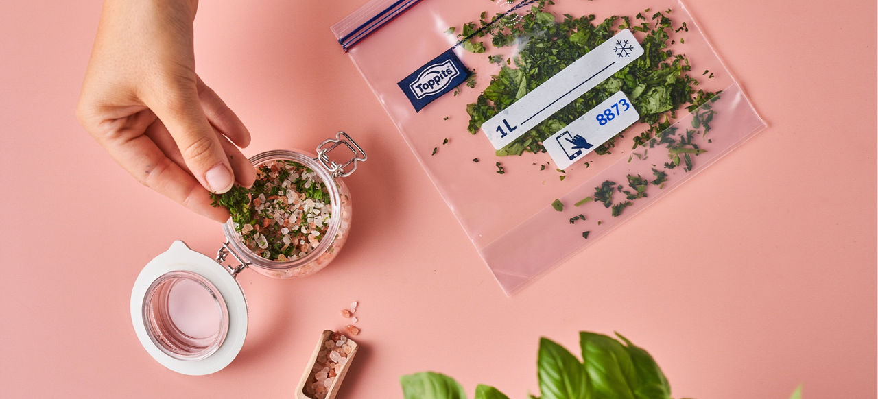 Hand gibt gehackte Kräuter aus einem Gefrierbeutel in ein kleines Glas auf einem rosa Untergrund. Neben dem Glas liegt ein Toppits Gefrierbeutel mit Kräutern. 