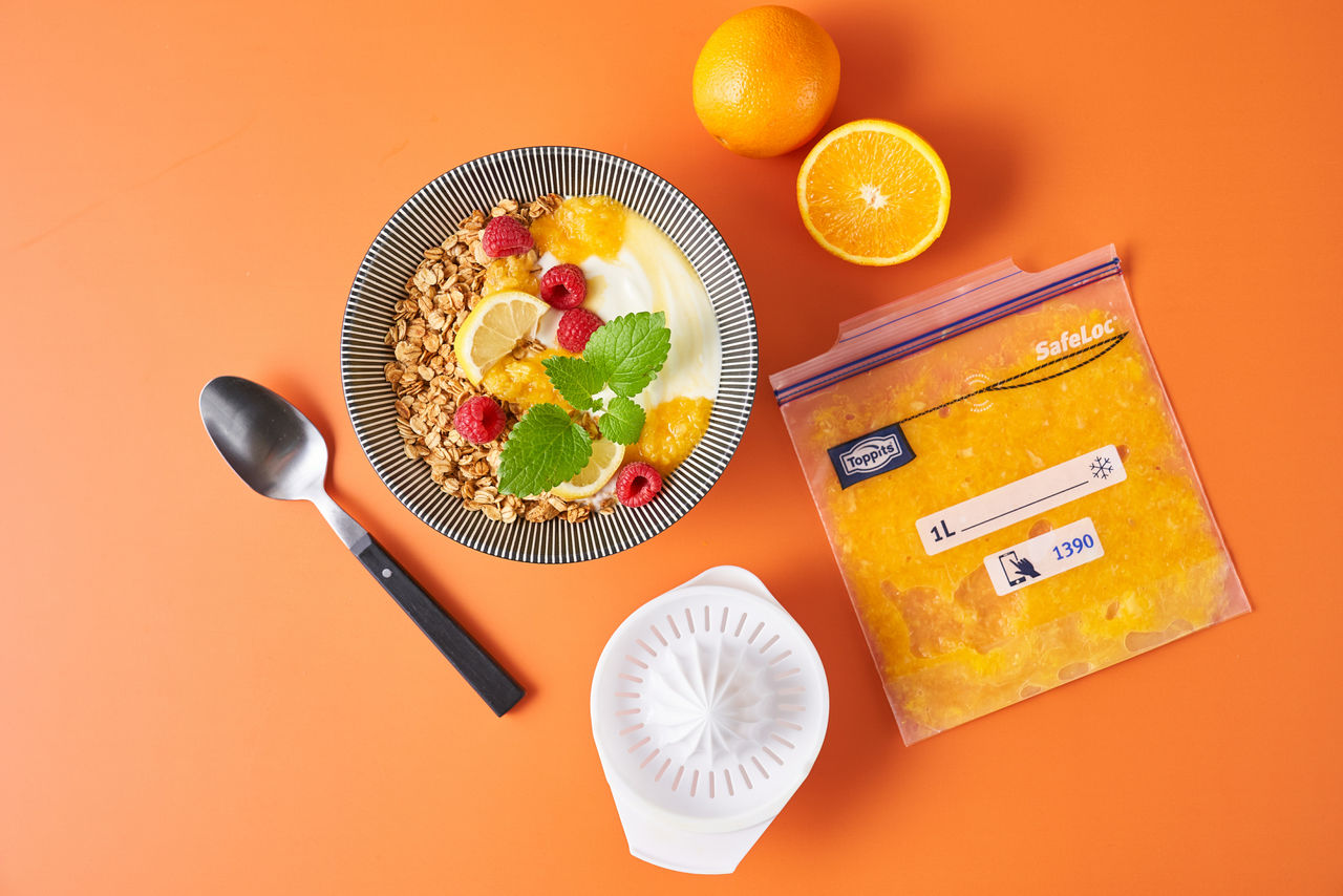 Schale mit Joghurt, Müsli, Himbeeren, Zitronenscheibe und Orangensaft auf einem orangefarbenen Hintergrund, daneben ein Toppits SafeLoc Gefrierbeutel mit gefrorenem Fruchtfleisch, eine halbe und eine ganze Orange sowie eine Zitronenpresse